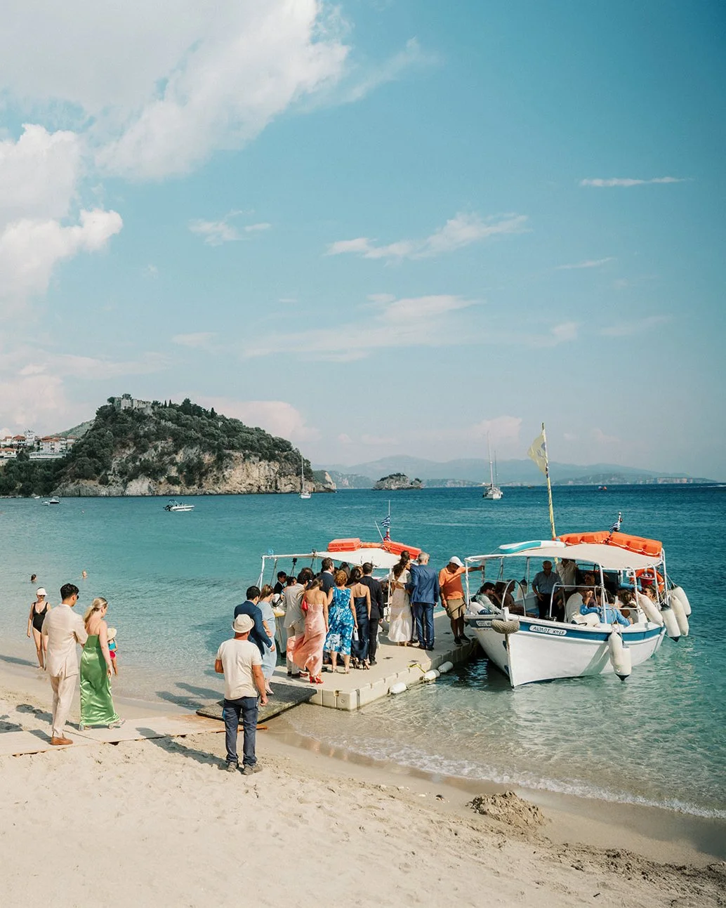 People preparing to board boats at a beach dock with a scenic island and calm sea in the background.