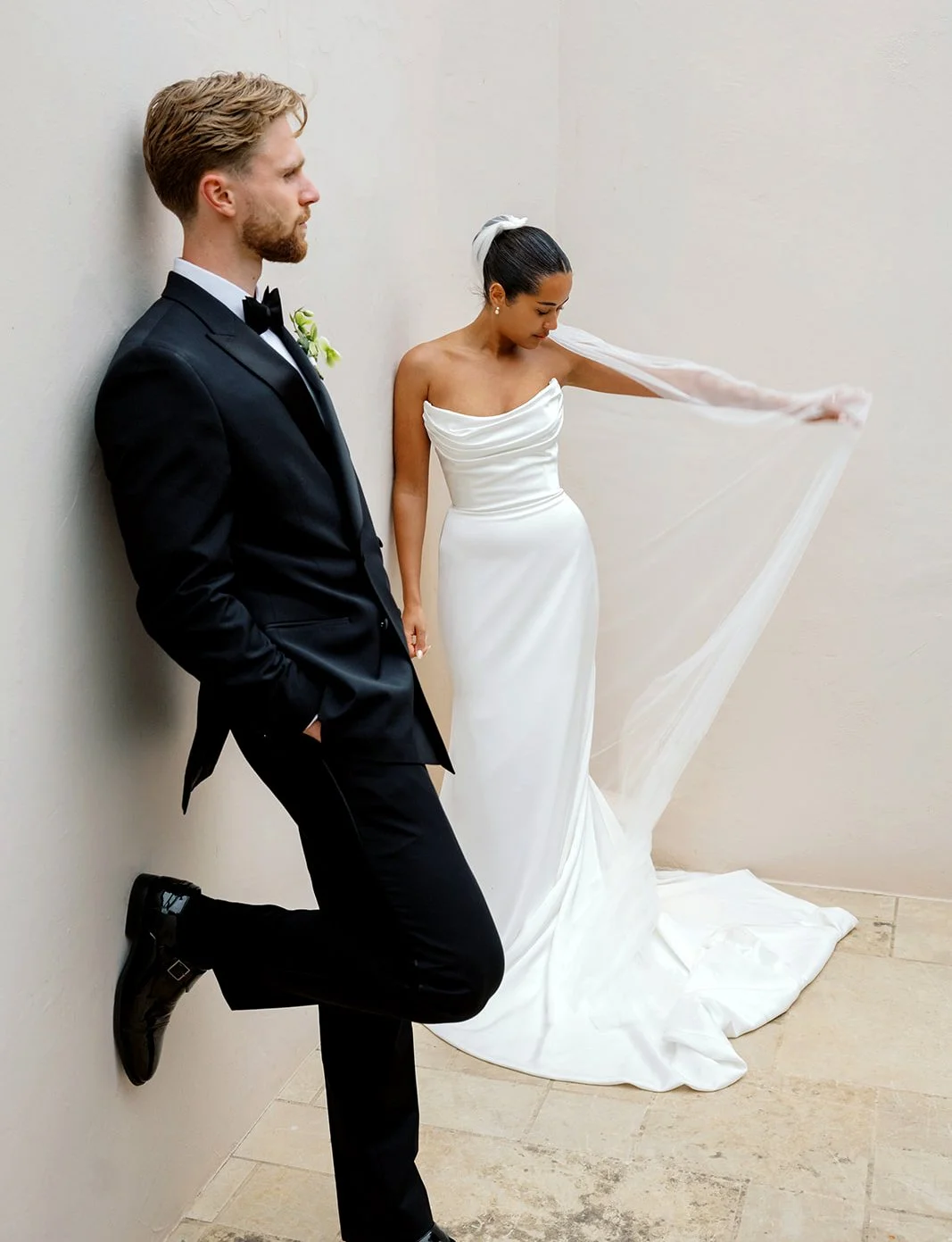 A bride in a white wedding dress and veil leaning against a wall, and a groom in a black tuxedo with a boutonniere, standing with one foot on the wall as he leans back, both in a modern indoor setting.
