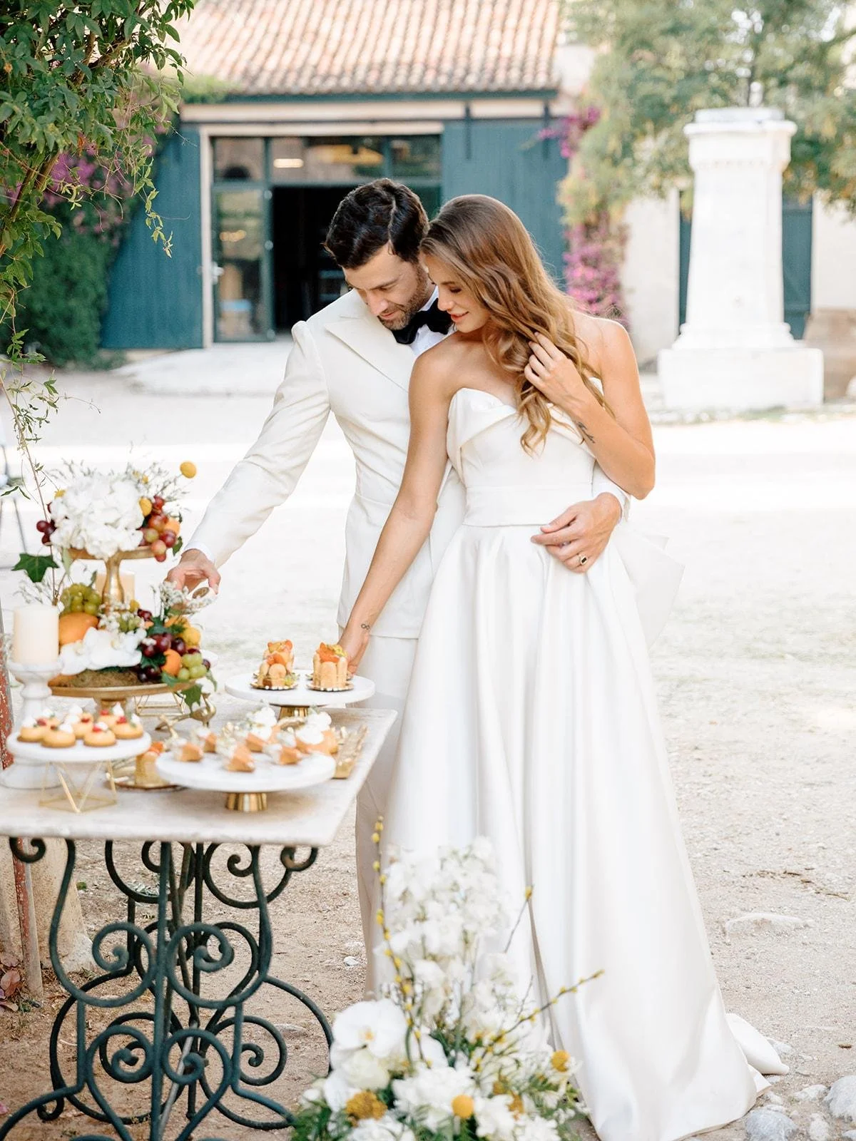 Bride and groom in white wedding attire sharing a moment near a dessert table outdoors with cupcakes and fruits.