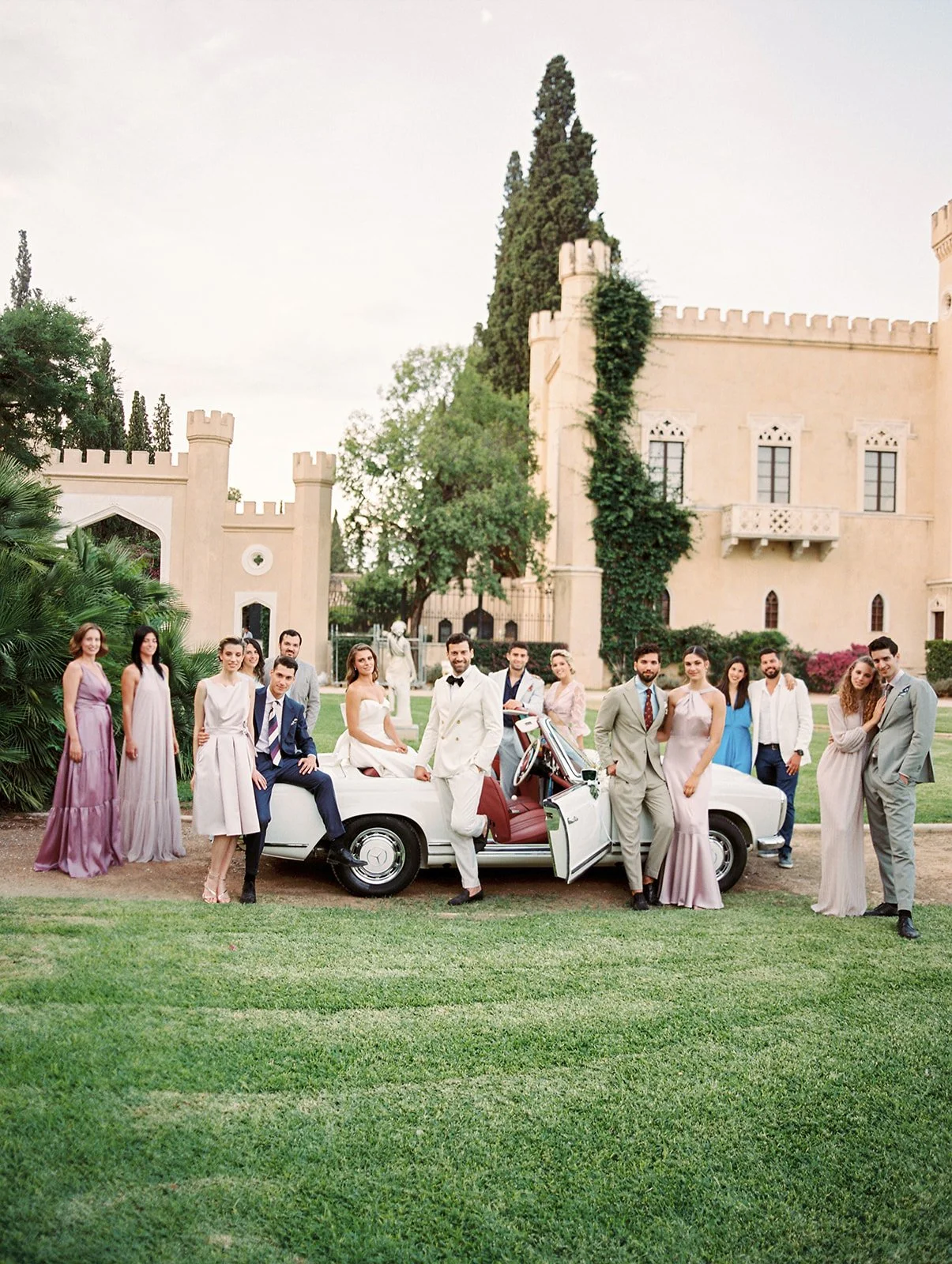 A group of people in formal attire gathers around a white convertible car in front of a historic castle-like building with greenery.