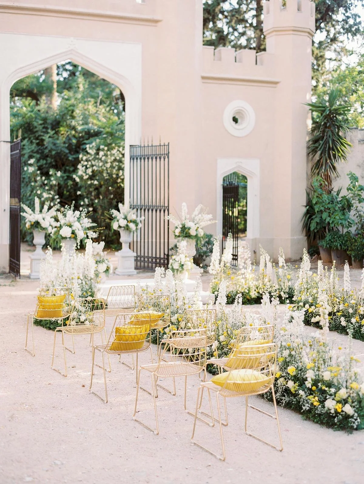 Outdoor wedding setup with yellow chairs and white floral arrangements in front of a castle-like structure.