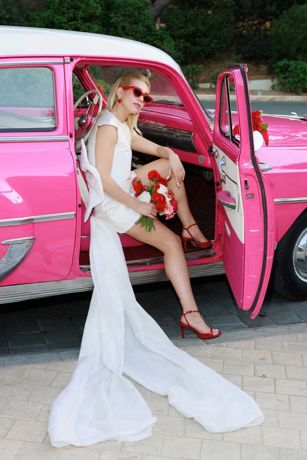 A woman in a white dress and red high heels sitting in a pink vintage car, holding a bouquet of red and pink flowers, wearing red sunglasses, with a white long train flowing from her dress.