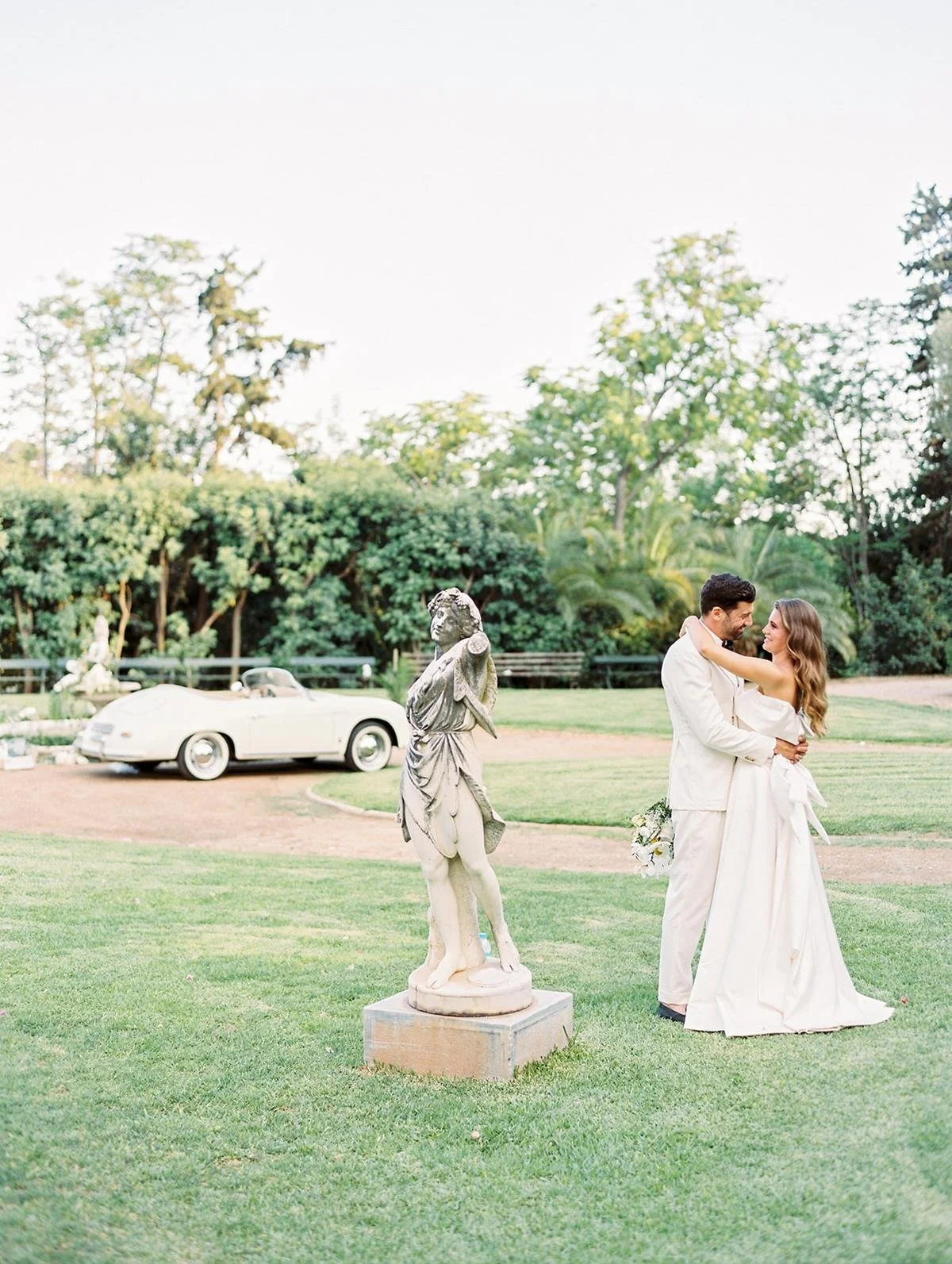A couple dressed in wedding attire embracing outdoors on a grassy area with trees in the background. There is a vintage car and classical sculpture nearby.