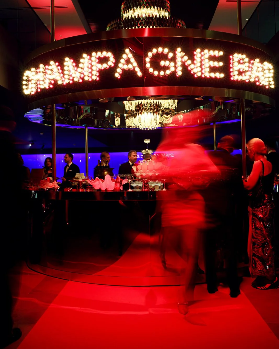 A bar with a rounded counter and a large illuminated sign that reads 'HANA PABEN' in bright lights. People in formal attire are socializing, and the bartender is preparing drinks. The setting is elegant with chandeliers and colorful lighting.