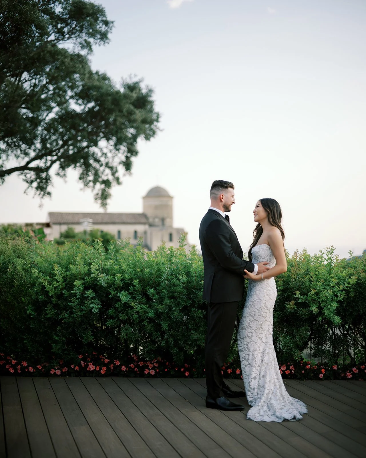A bride and groom standing face-to-face on a wooden platform, holding hands, with a large bush and flowers behind them. The bride is wearing a white lace wedding gown, and the groom is in a black tuxedo. A historic building is visible in the backgrou