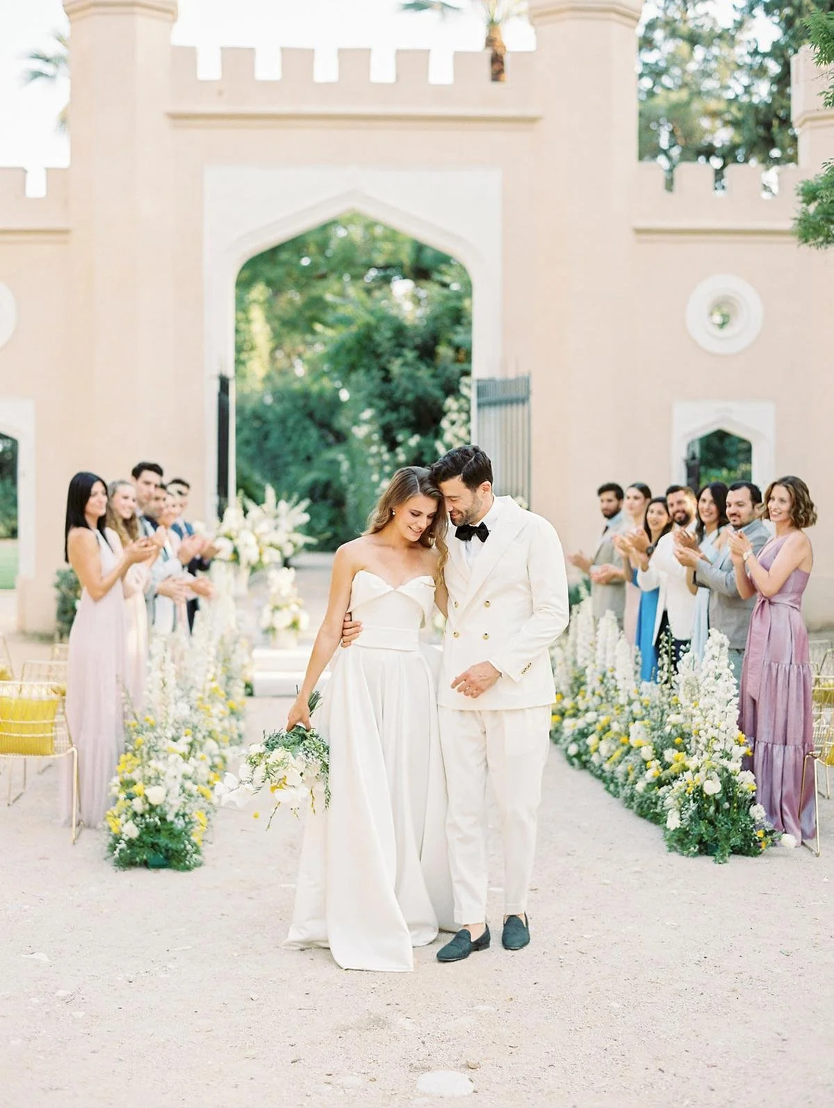A bride and groom walking together at their wedding ceremony, surrounded by friends and family, with floral decorations and a historic pink castle in the background.