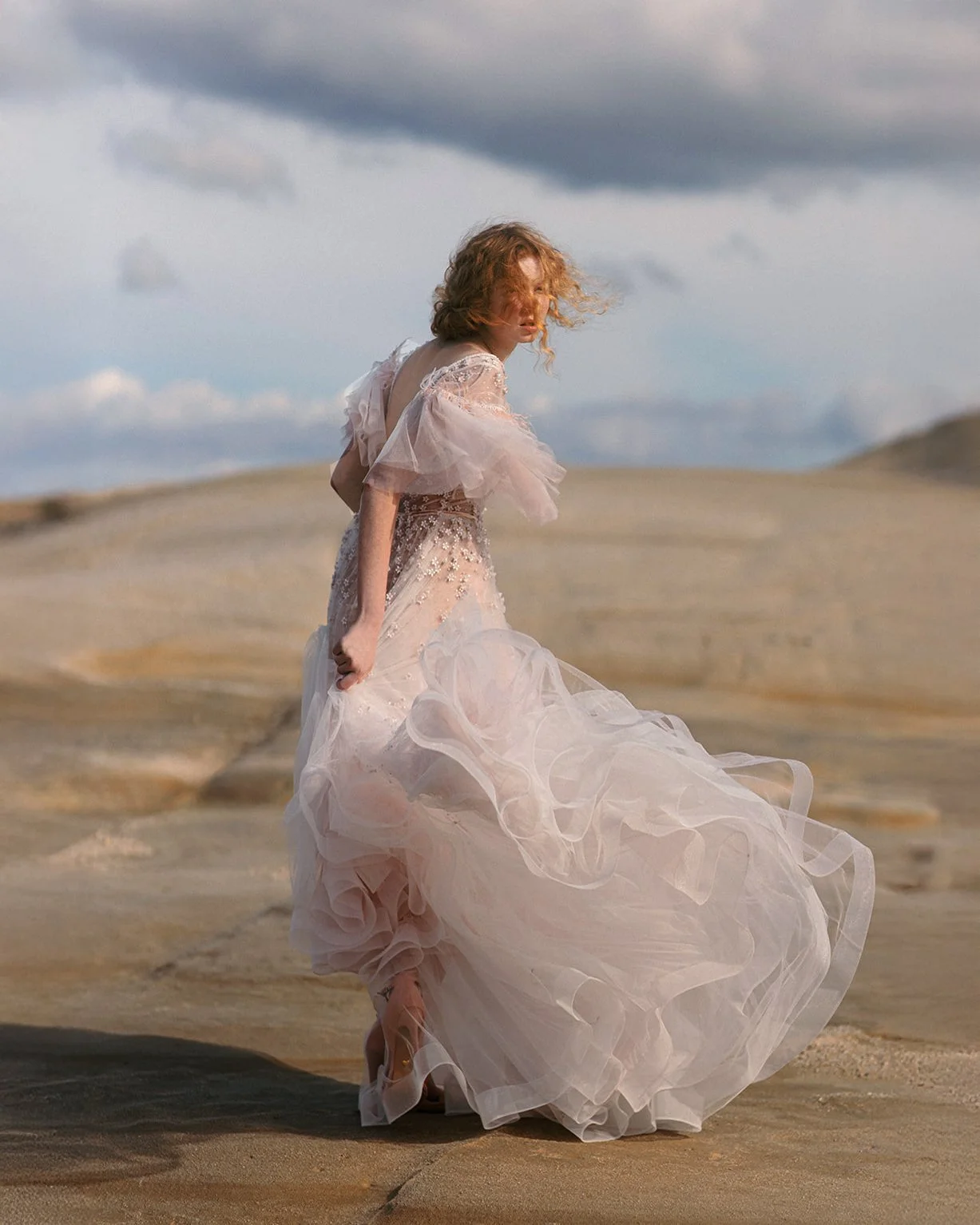 A woman in a flowing, light pink, ruffled dress standing on a sandy landscape with cloudy skies in the background.