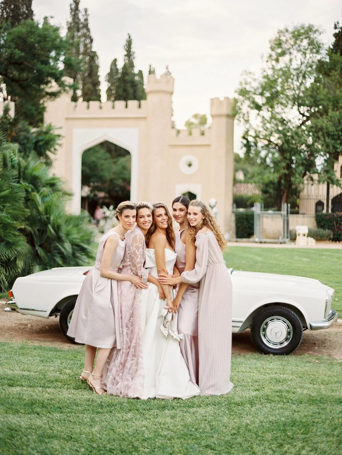 Group of five women in elegant dresses standing by a vintage white car outdoors with a castle-like structure in the background.