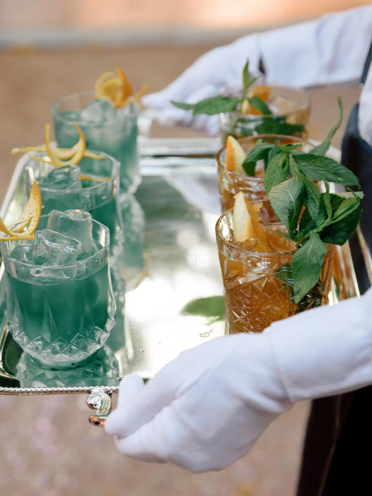 A silver tray carrying several cocktails in crystal glasses, garnished with lemon wedges and fresh mint leaves.