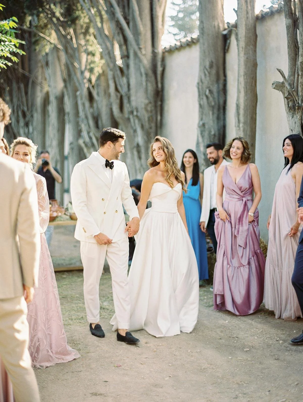 A wedding couple walking hand in hand outdoors, surrounded by guests, with trees and a rustic wall in the background.