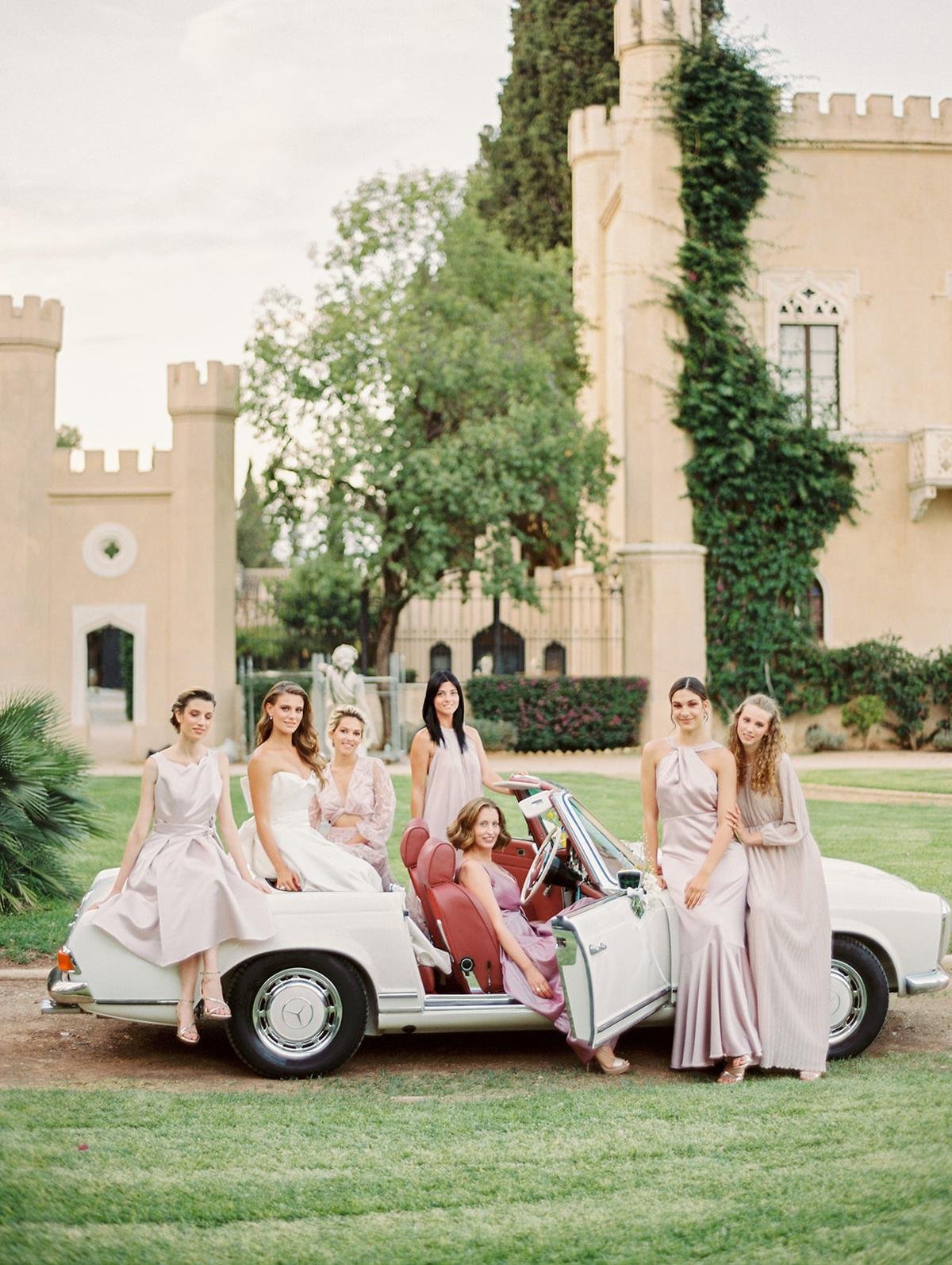 Group of women in elegant dresses posing with a vintage convertible car in front of a castle-like building with lush greenery.