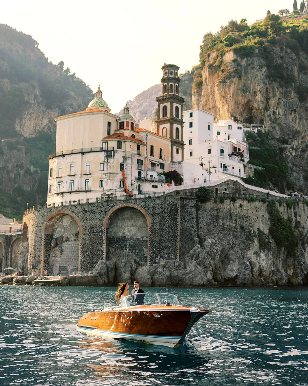 A couple in wedding attire on a wooden speedboat in water with a coastal town built on a rocky hillside with historic buildings and lush greenery in the background.