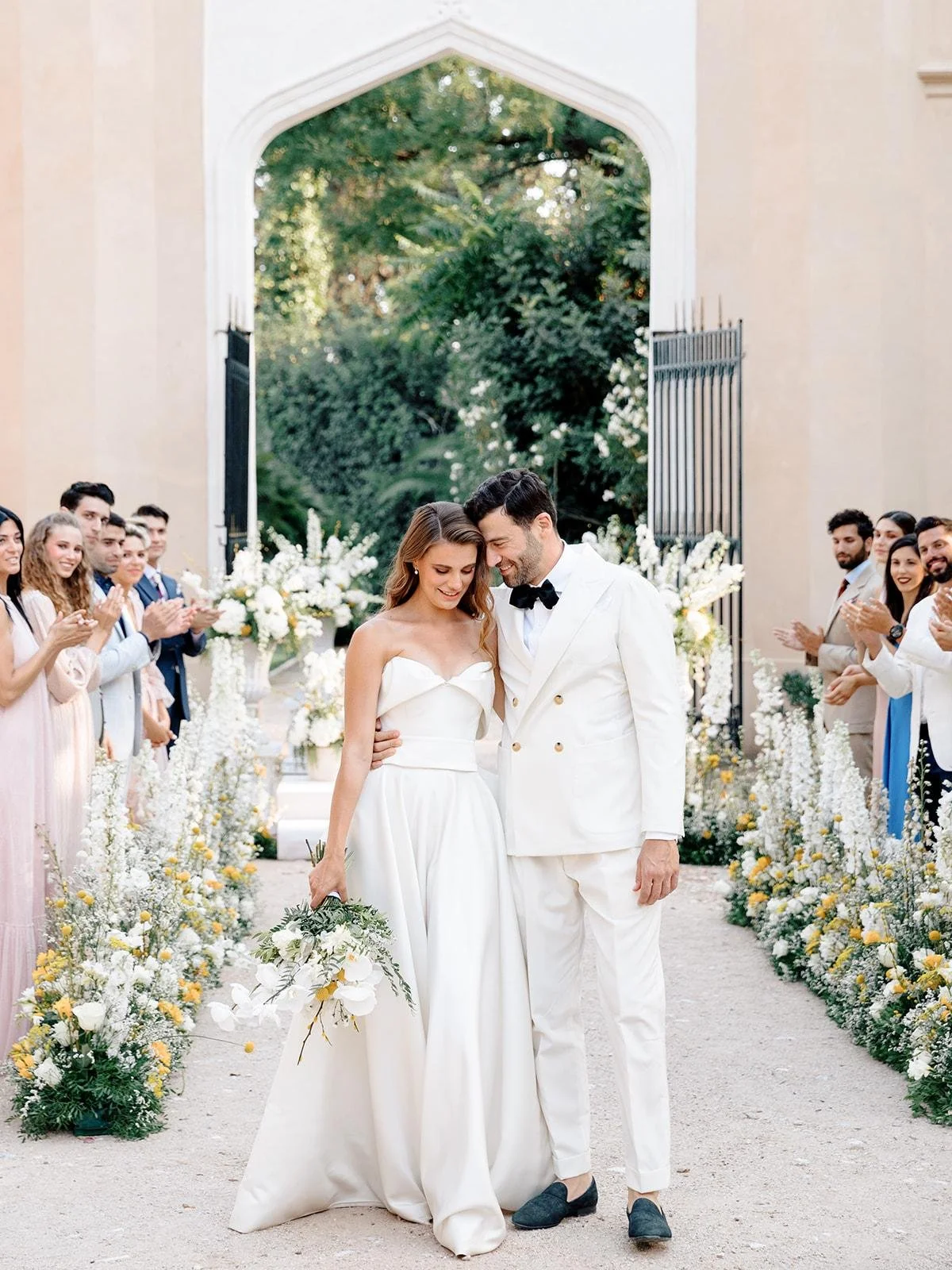A bride and groom in wedding attire walking arm in arm through a floral aisle with guests applauding on both sides during an outdoor wedding ceremony.