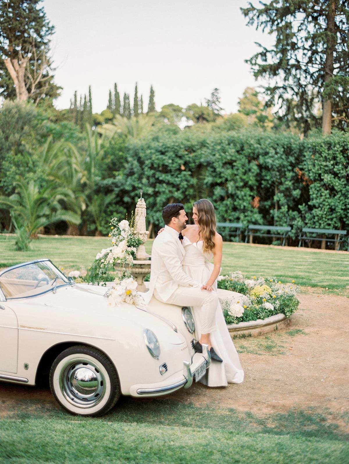 A couple in wedding attire sitting on the hood of a vintage white convertible car, surrounded by flowers and garden greenery.