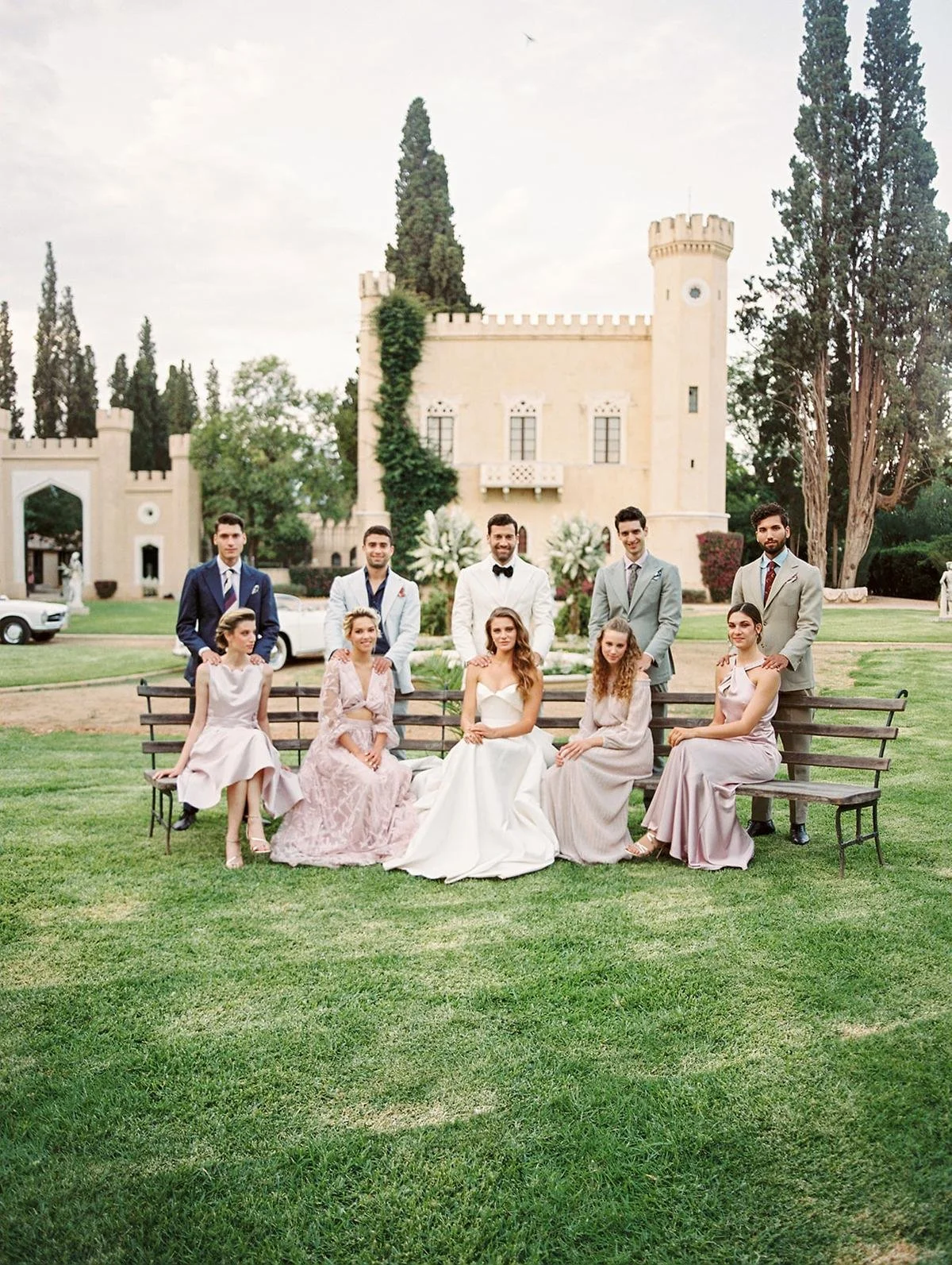 Group of people in formal attire posing on a lawn with a castle-like building and vintage cars in the background.