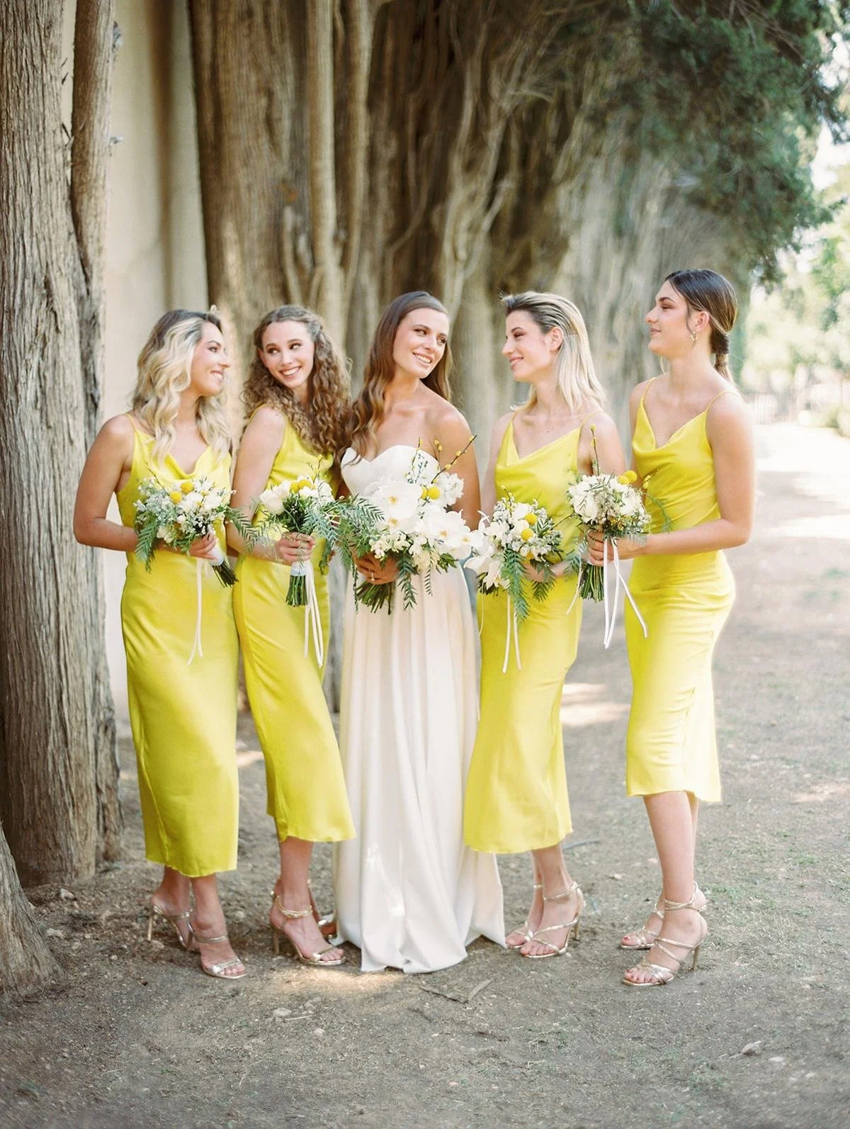 A bride and four bridesmaids standing outdoors by tall trees, smiling and holding bouquets of white and yellow flowers. The bride wears a white dress; the bridesmaids wear yellow dresses.