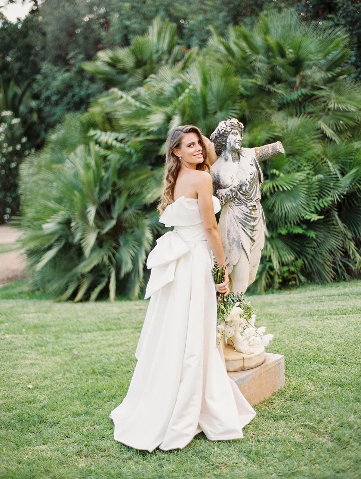 A woman in a white strapless wedding dress holding a bouquet, standing beside a classical statue in a garden with lush green tropical plants.