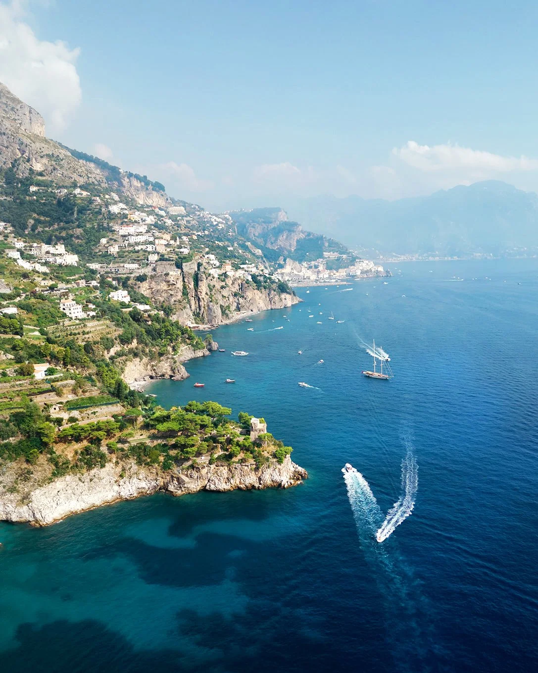 Coastal landscape with cliffs, green vegetation, and white buildings, along with boats and yachts on the blue water, under a partly cloudy sky.