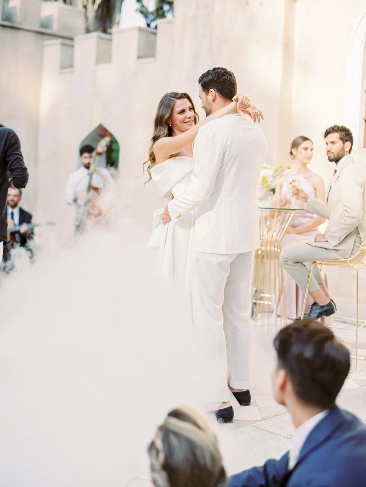 A couple dancing at a wedding reception with the bride wearing a white dress and the groom in a white suit. Guests are seated nearby, some watching, with a musician in the background playing a guitar. The setting appears to be a bright indoor or outd