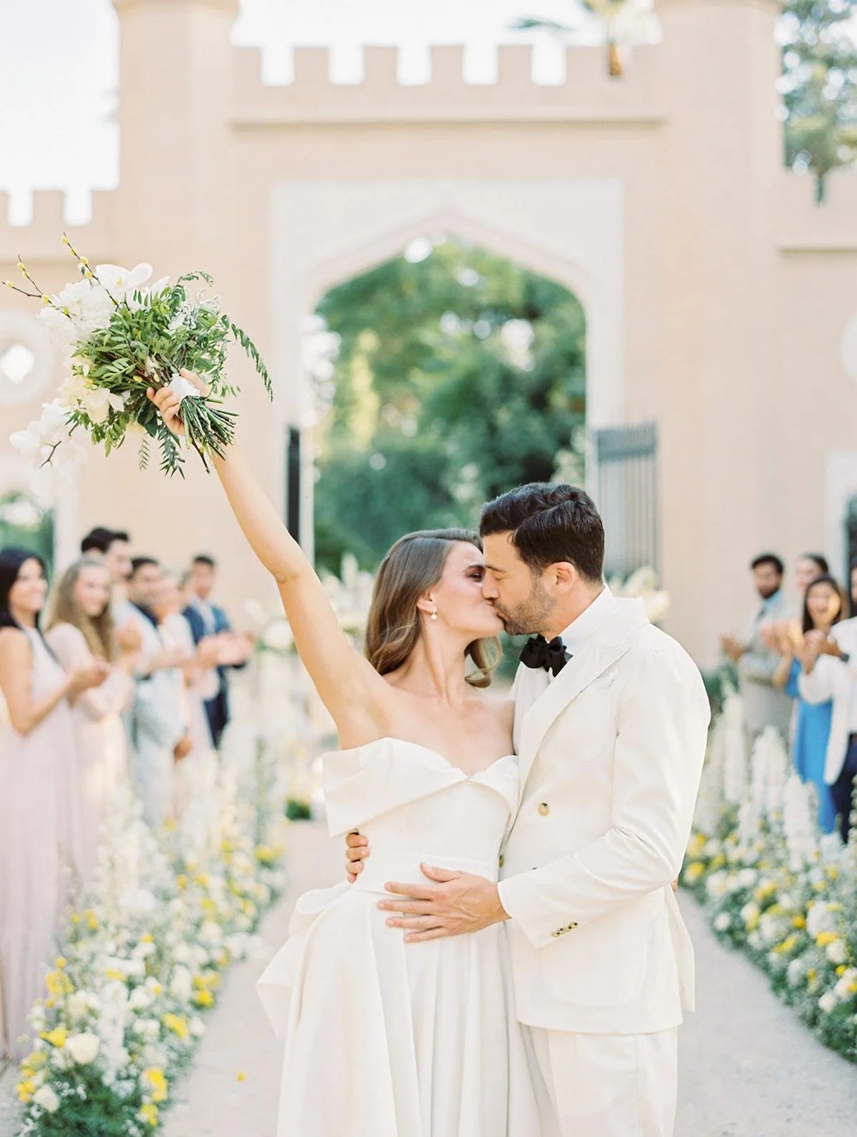 A newlywed couple sharing a kiss at their outdoor wedding ceremony, with the bride holding a bouquet of flowers in the air.