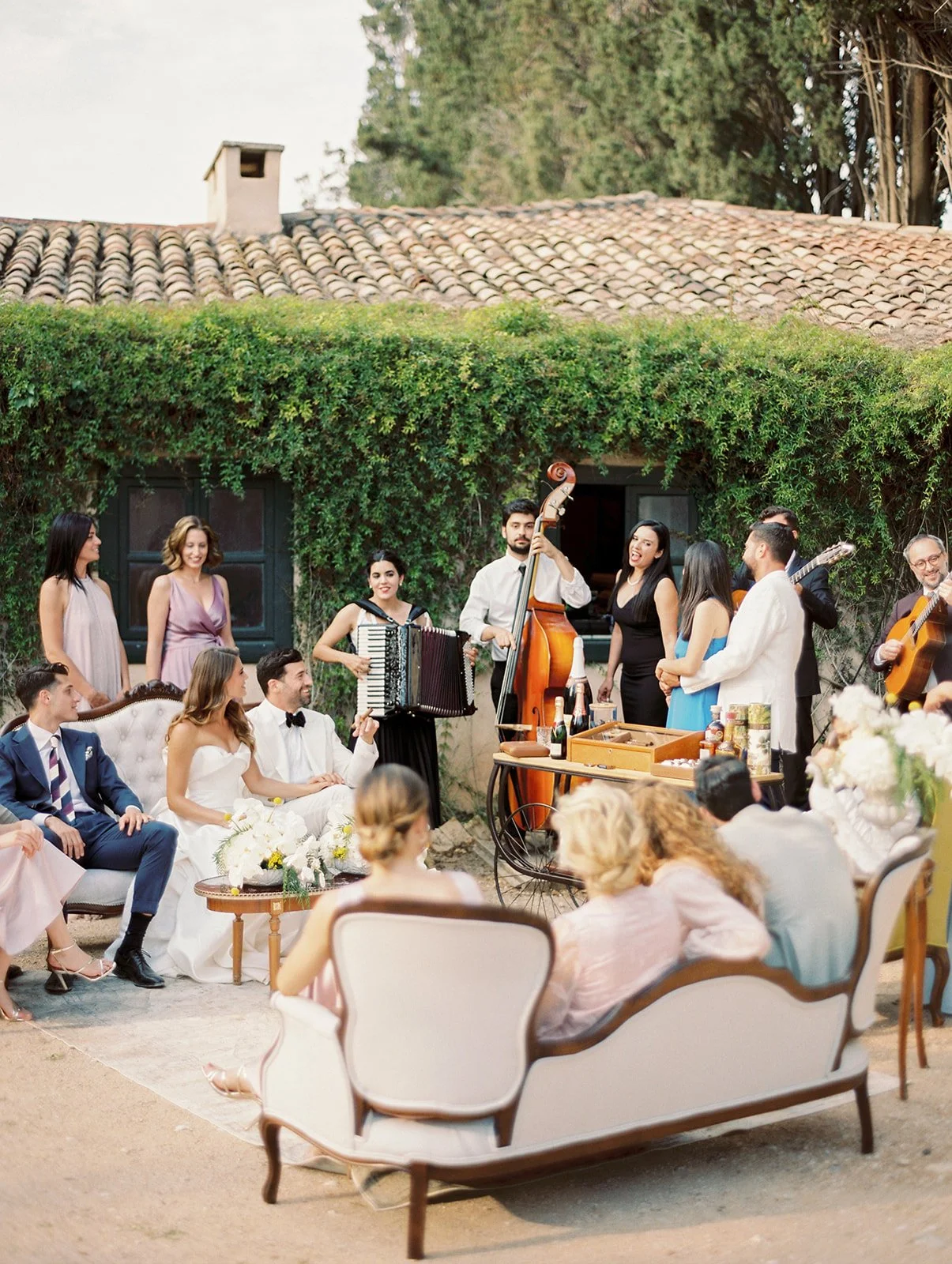 A wedding reception outdoors with guests seated and a live band performing, featuring women and men in formal attire near a rustic house with a tile roof.
