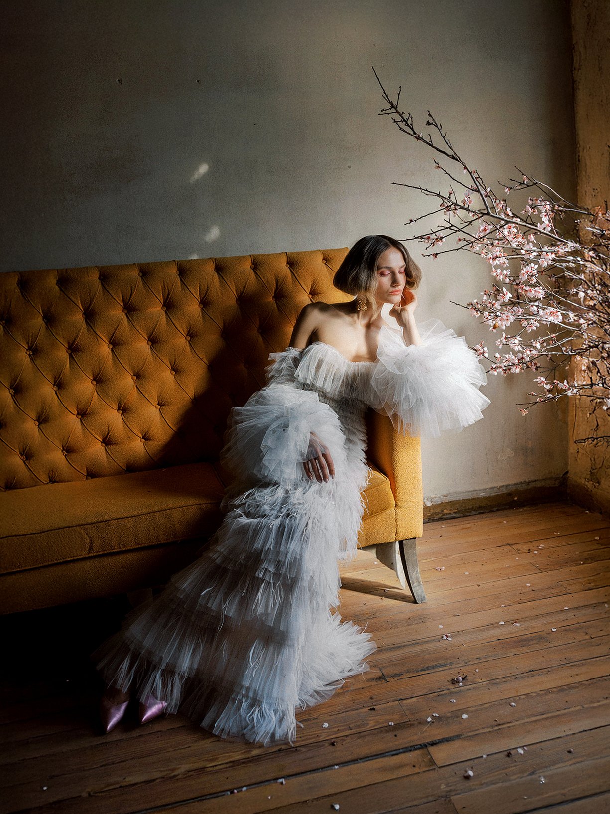 A woman in a white ruffled gown sitting on a mustard-colored vintage sofa in a room with wooden floors and peeling walls, near a branch of pink blossoms.