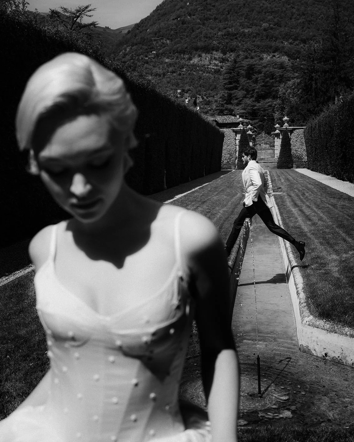 Black and white photograph of a woman in a wedding dress in the foreground and a man in a tuxedo crossing a small water channel in the background, with a pathway and mountain landscape behind.