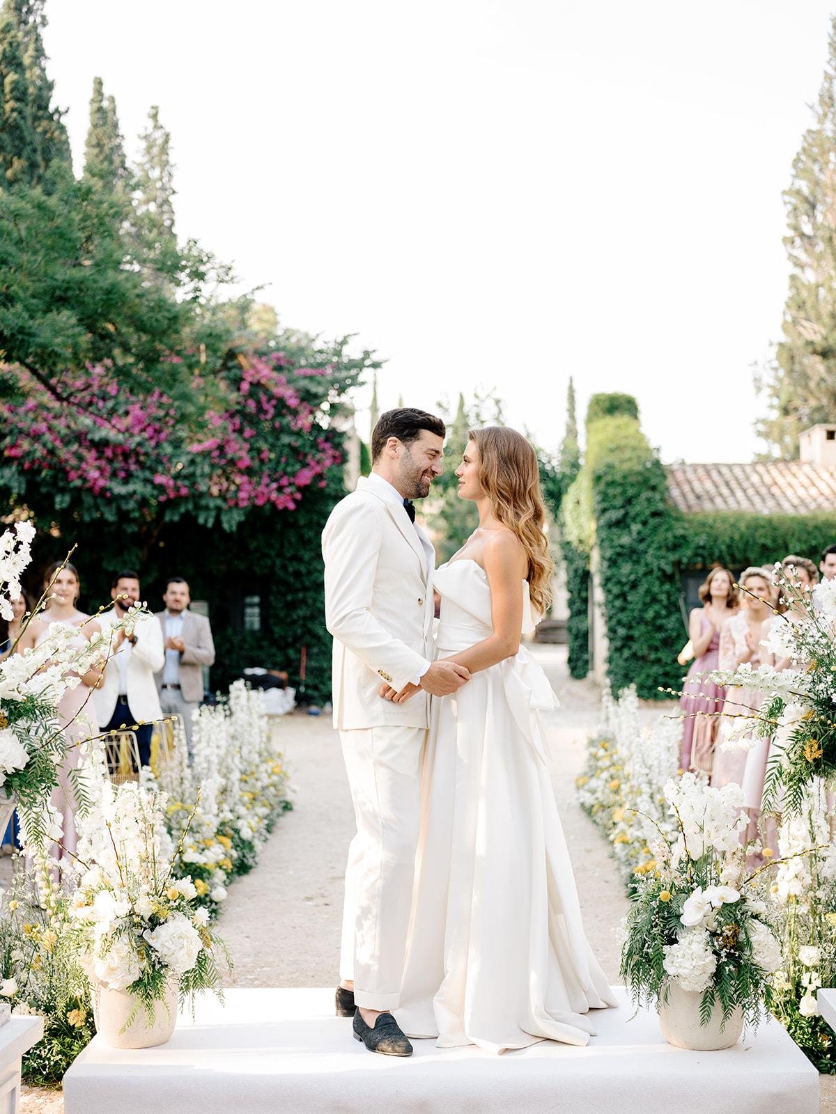 A couple in wedding attire stands on a platform during their outdoor wedding ceremony, holding hands and smiling at each other, surrounded by friends, family, and floral arrangements.