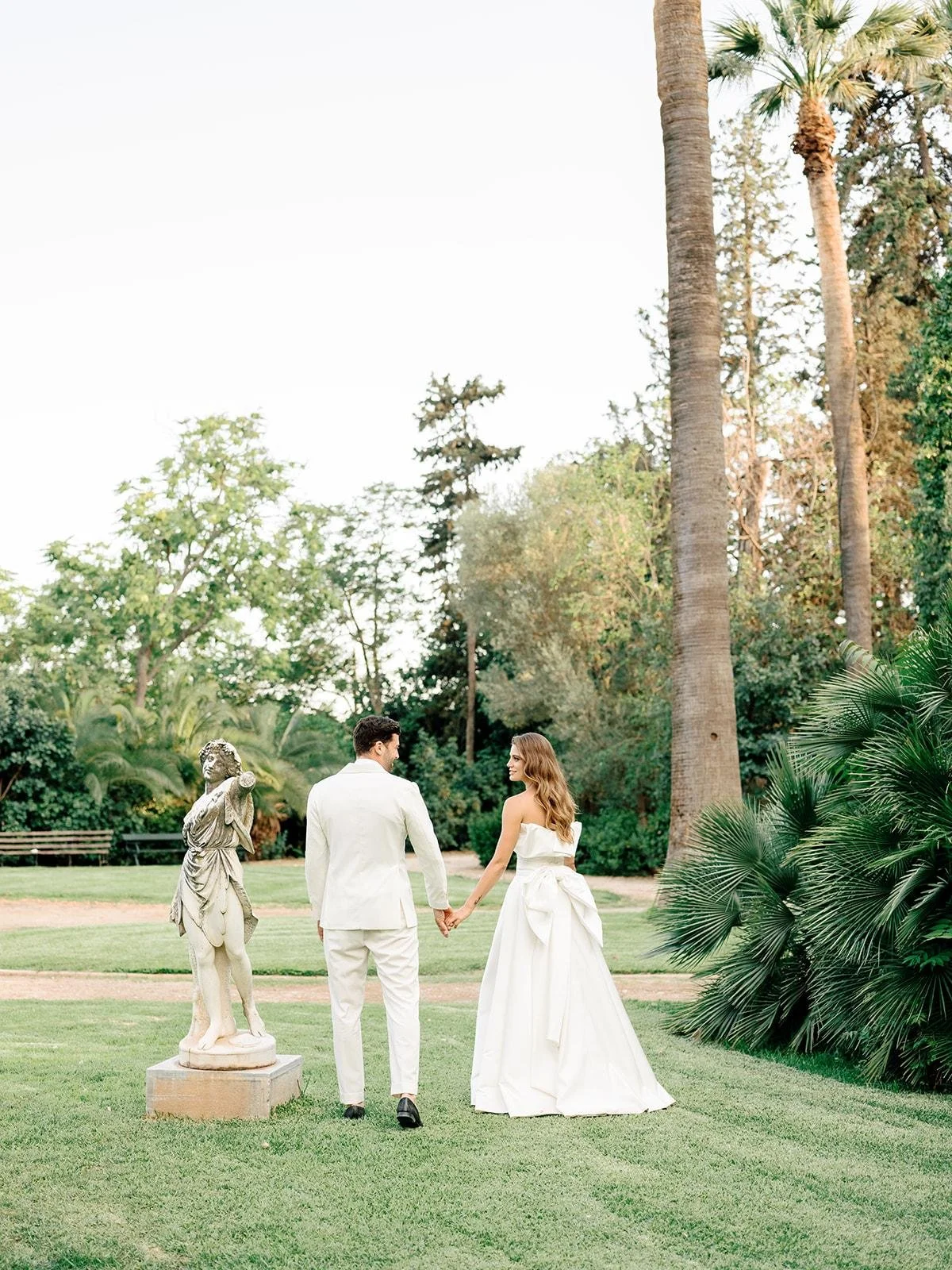 A couple dressed in wedding attire holding hands and walking in a lush garden with tall palm trees and greenery.