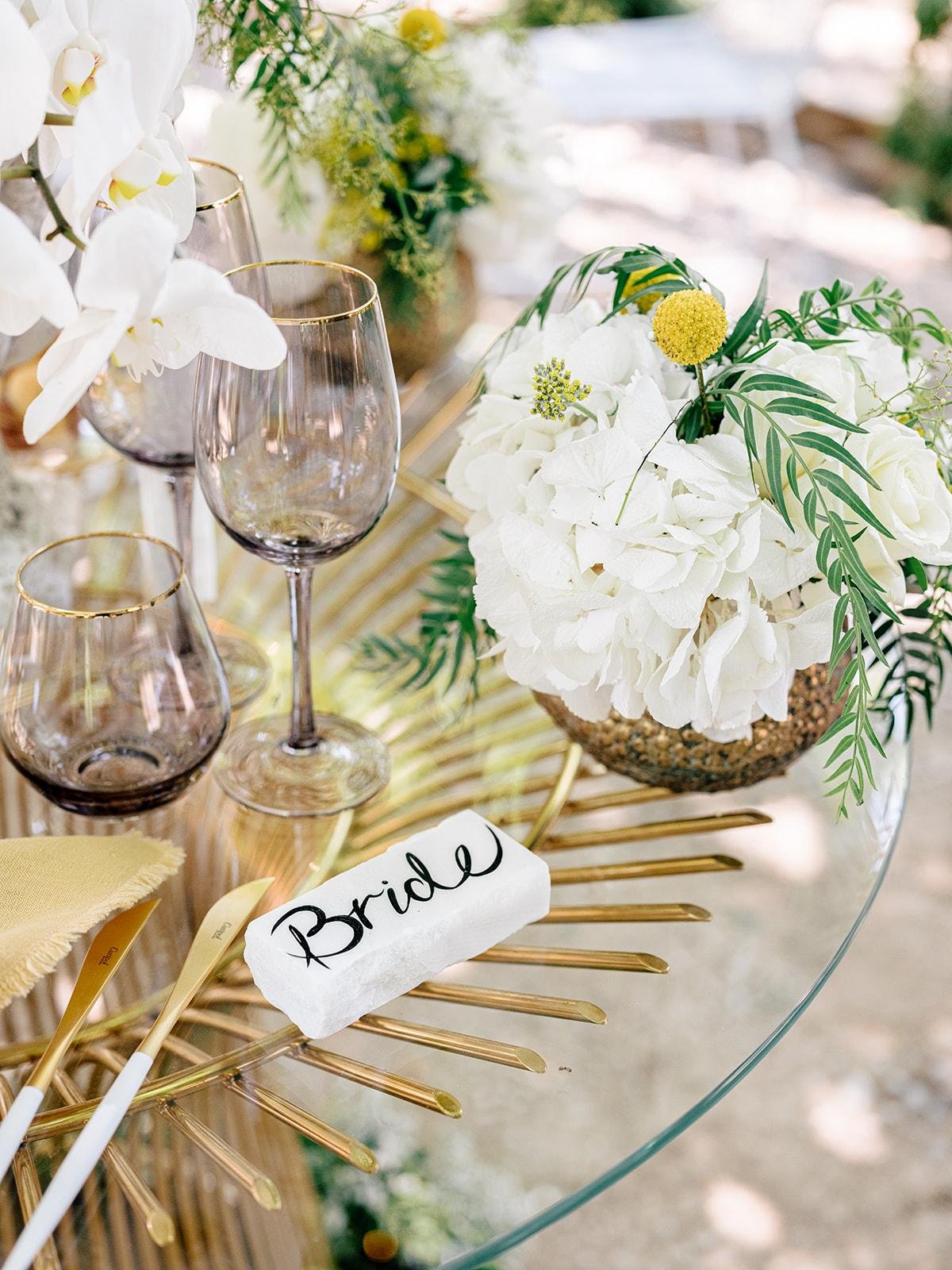 Wedding table with a floral arrangement of white flowers and greenery, three purple wine glasses, and a place card labeled 'Bride' set on a glass table.