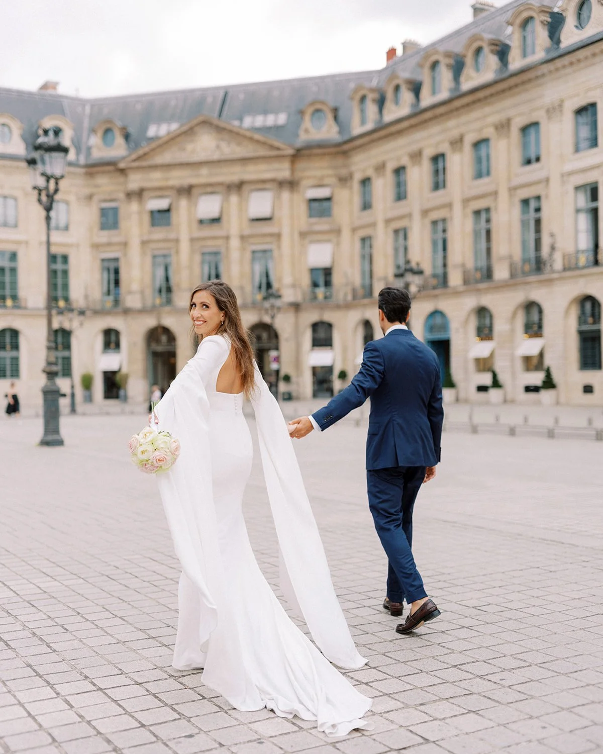 Bride in a white wedding gown holding a bouquet, smiling and looking back, while groom in a blue suit pulls her by hand in a European city square with historic buildings and cobblestone pavement.