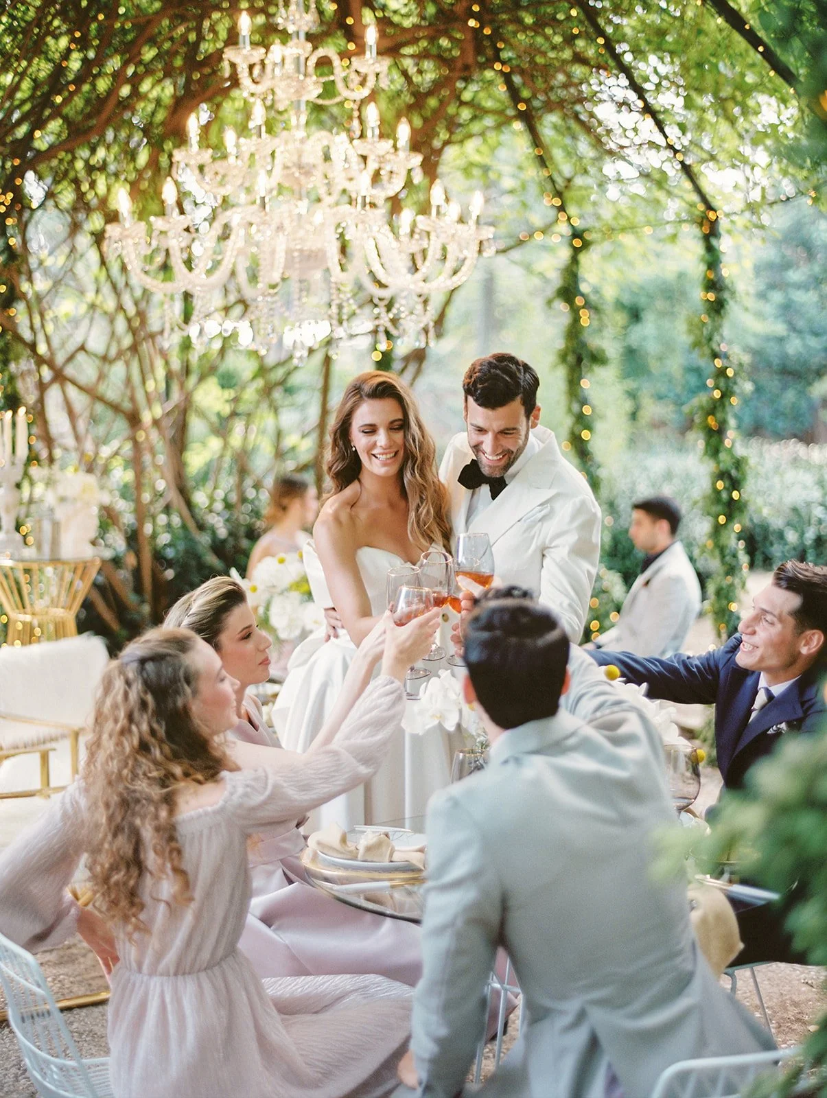 People celebrating at a wedding reception outdoors with a chandelier and string lights, raising glasses in a toast.