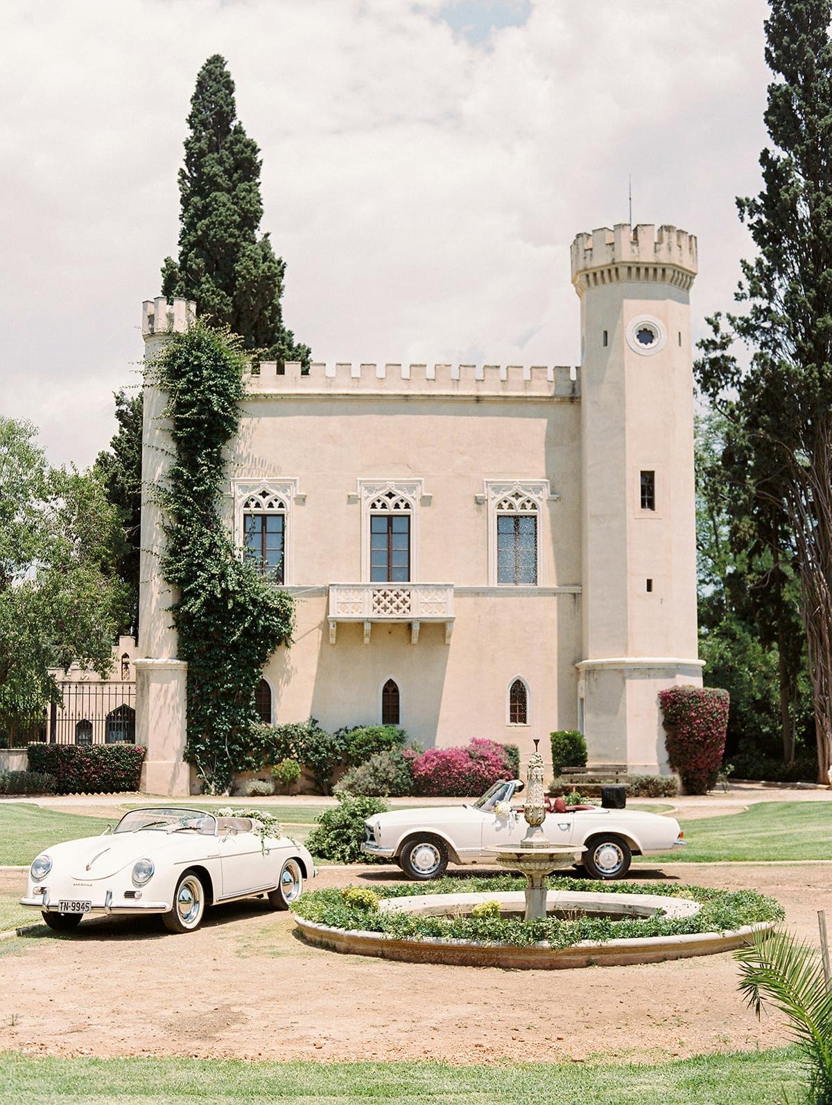 A white castle-like building with battlements, a tower, arched windows, surrounded by greenery and pink bushes, with two white vintage cars parked in front and a fountain with decorative urns in the foreground.