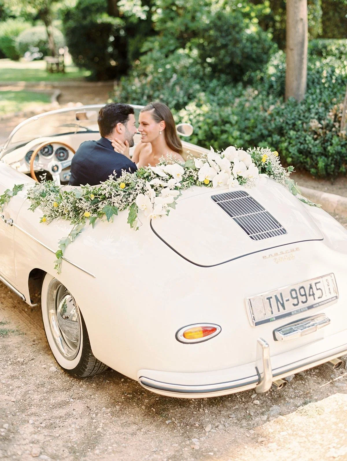 A bride and groom sitting in a white vintage convertible car decorated with a large floral arrangement, sharing a moment together on a wedding day.