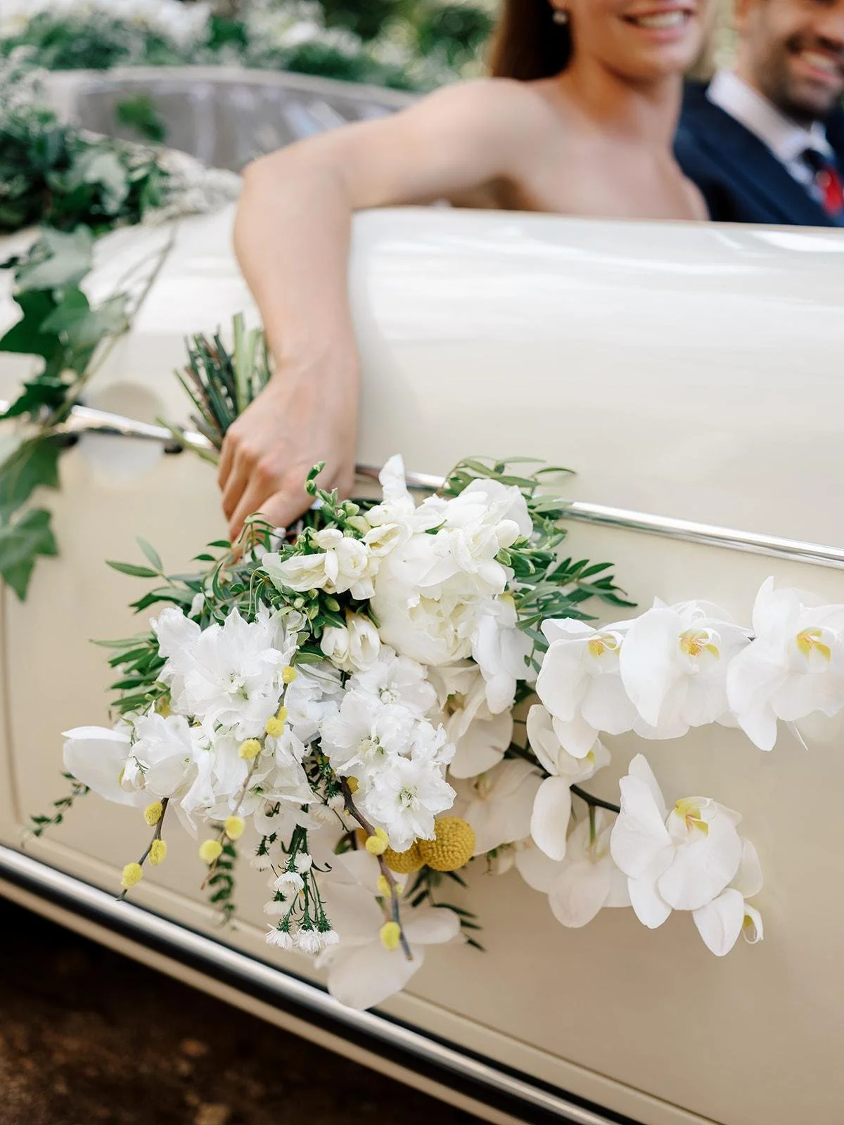 Bride and groom sitting in a vintage white car decorated with white and yellow flowers, with greenery and trees in the background.