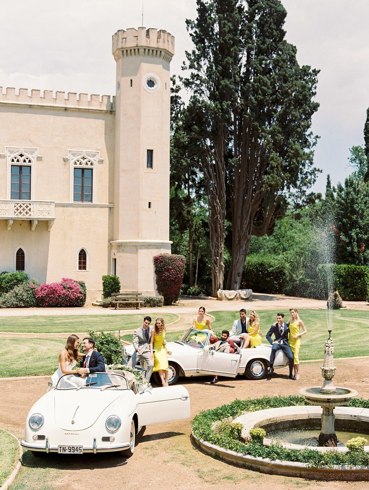 Group of people dressed in formal and semi-formal attire posing around vintage white cars in front of a large house with a fountain and lush greenery.