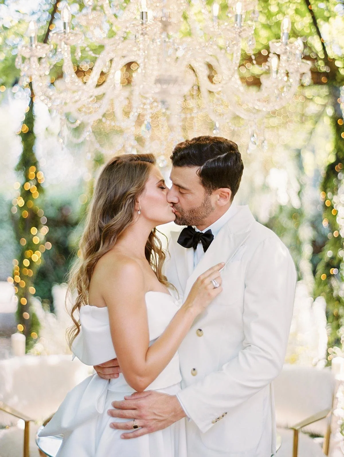 A romantic couple kissing at their wedding reception outdoors, with a chandelier hanging above and blurred string lights and greenery in the background.