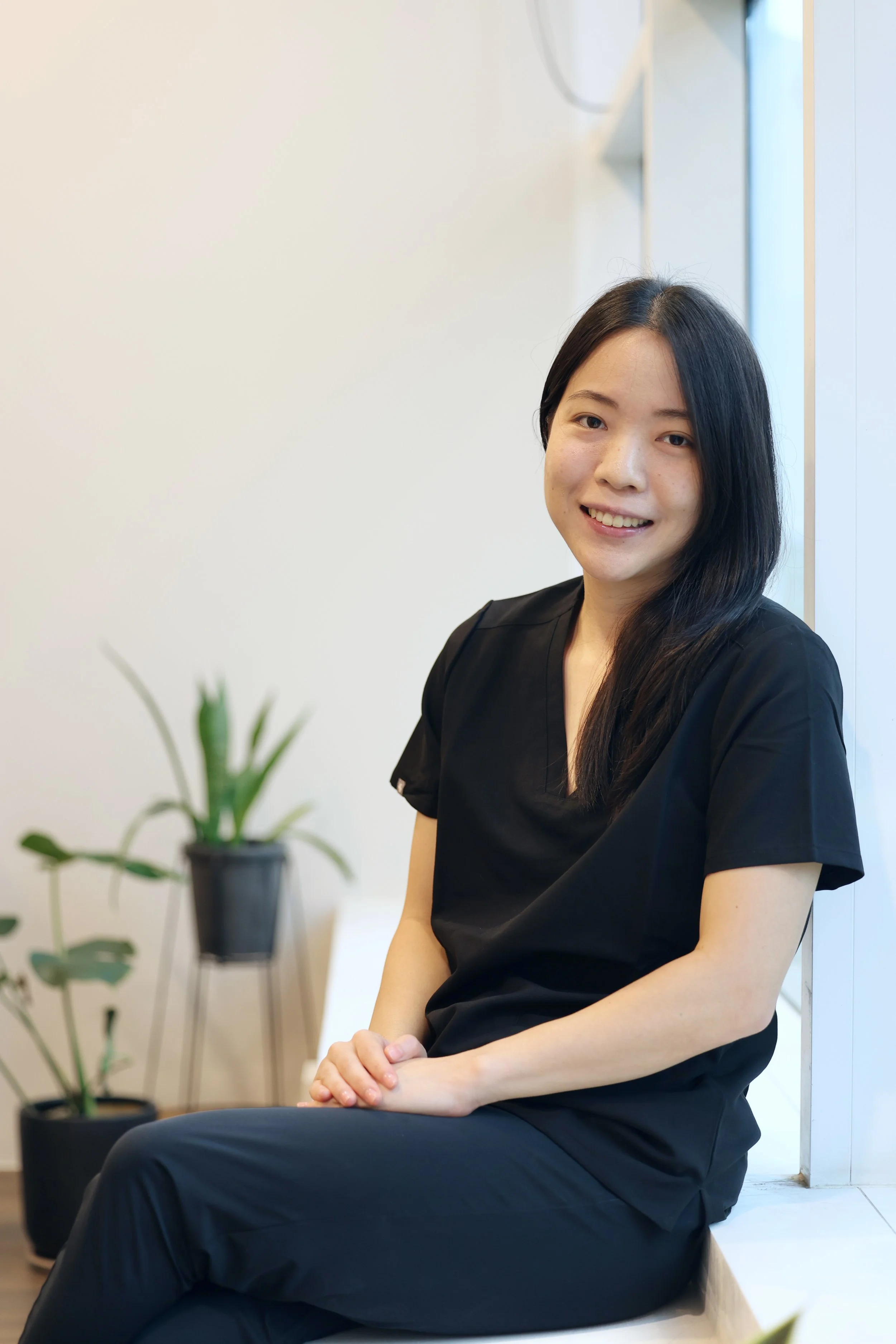A woman with long black hair wearing black medical scrubs sitting on a white counter, smiling in a bright room with white walls and a window. There is a potted plant in the foreground.