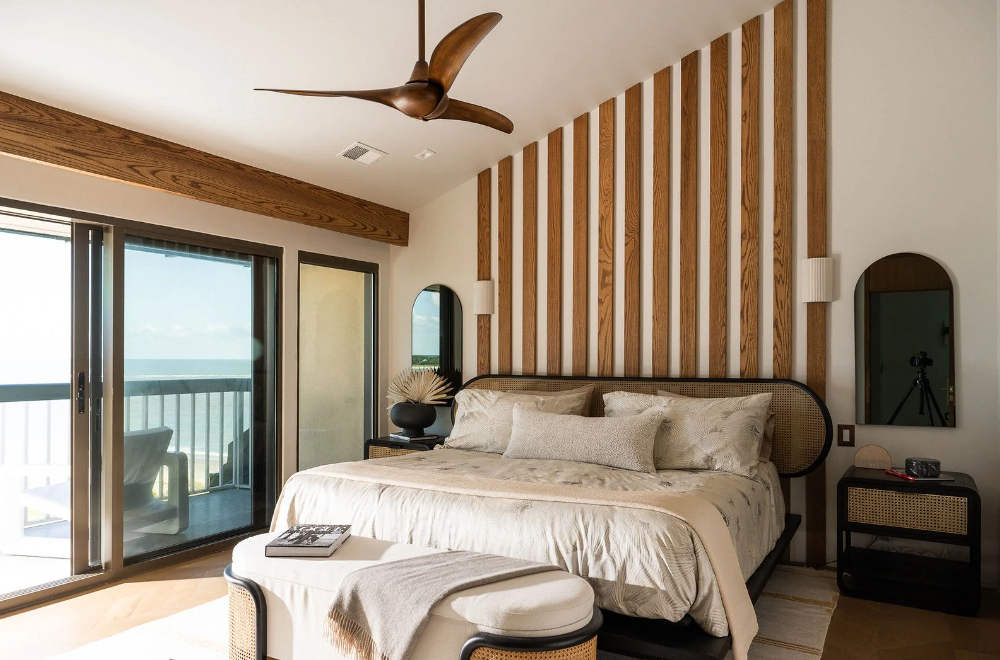 A bedroom with a large bed, beige bedding, and decorative pillows, wooden accents on the wall, a round mirror, a black nightstand, a clock, and a sliding glass door leading to a balcony with ocean view.