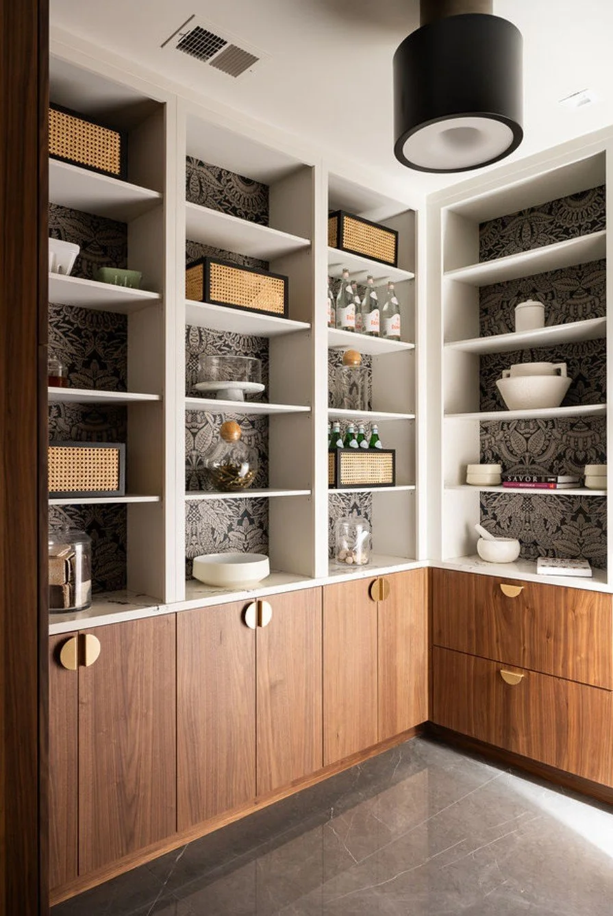 A corner of a room with white open shelves filled with decorative bowls, bottles, jars, and storage boxes. The lower cabinets are made of wood, and the back wall has dark patterned wallpaper. A black ceiling light is visible.