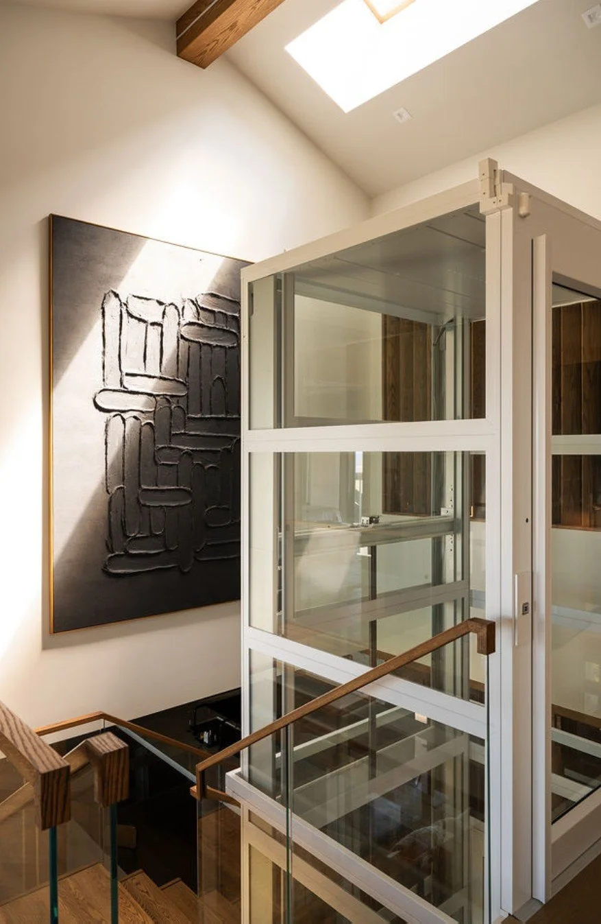 Modern interior with a glass elevator, a black textured artwork on the wall, a wooden staircase with glass railing, and a skylight ceiling.