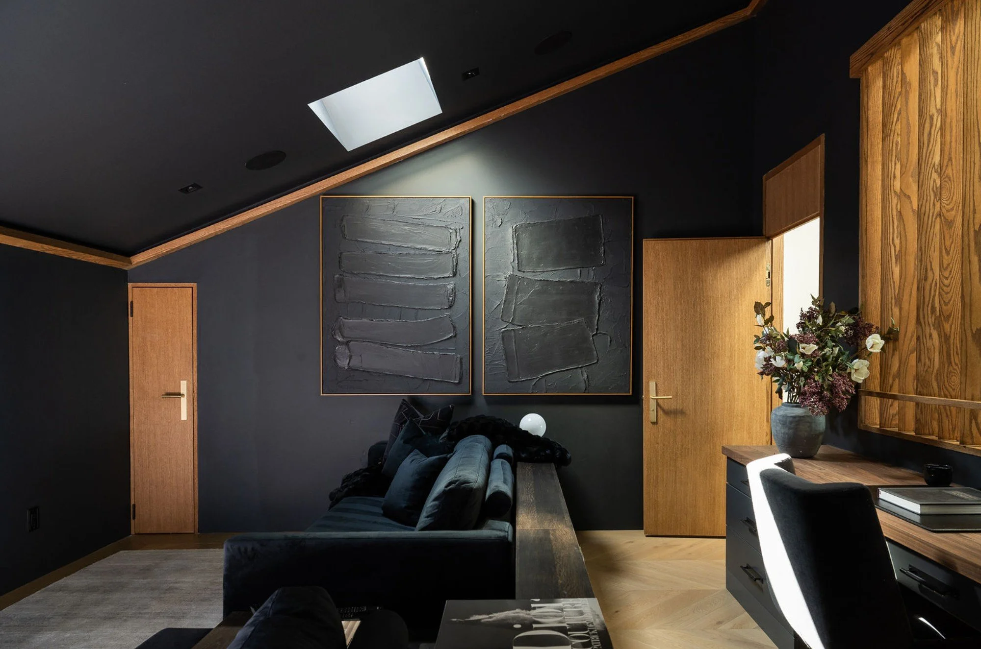 Modern living room with dark walls, wooden doors, and contemporary artwork on the wall. There is a dark gray sofa with black and gray pillows, a large gray vase with flowers, and a black entertainment unit with books.