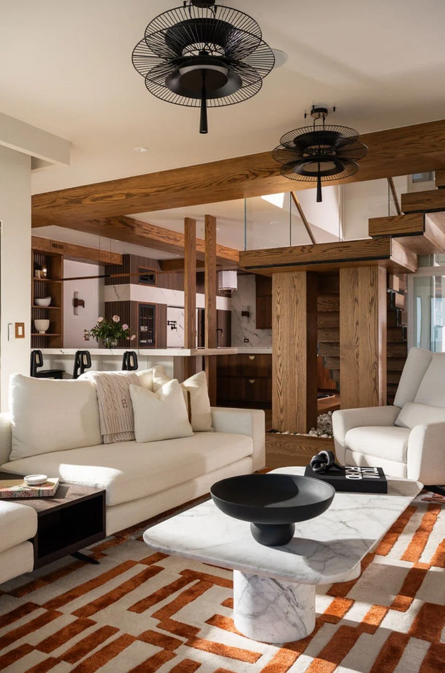 Living room with white sofa and armchair, marble coffee table with black bowl, wooden staircase, wood-paneled ceiling, and modern black ceiling fans.