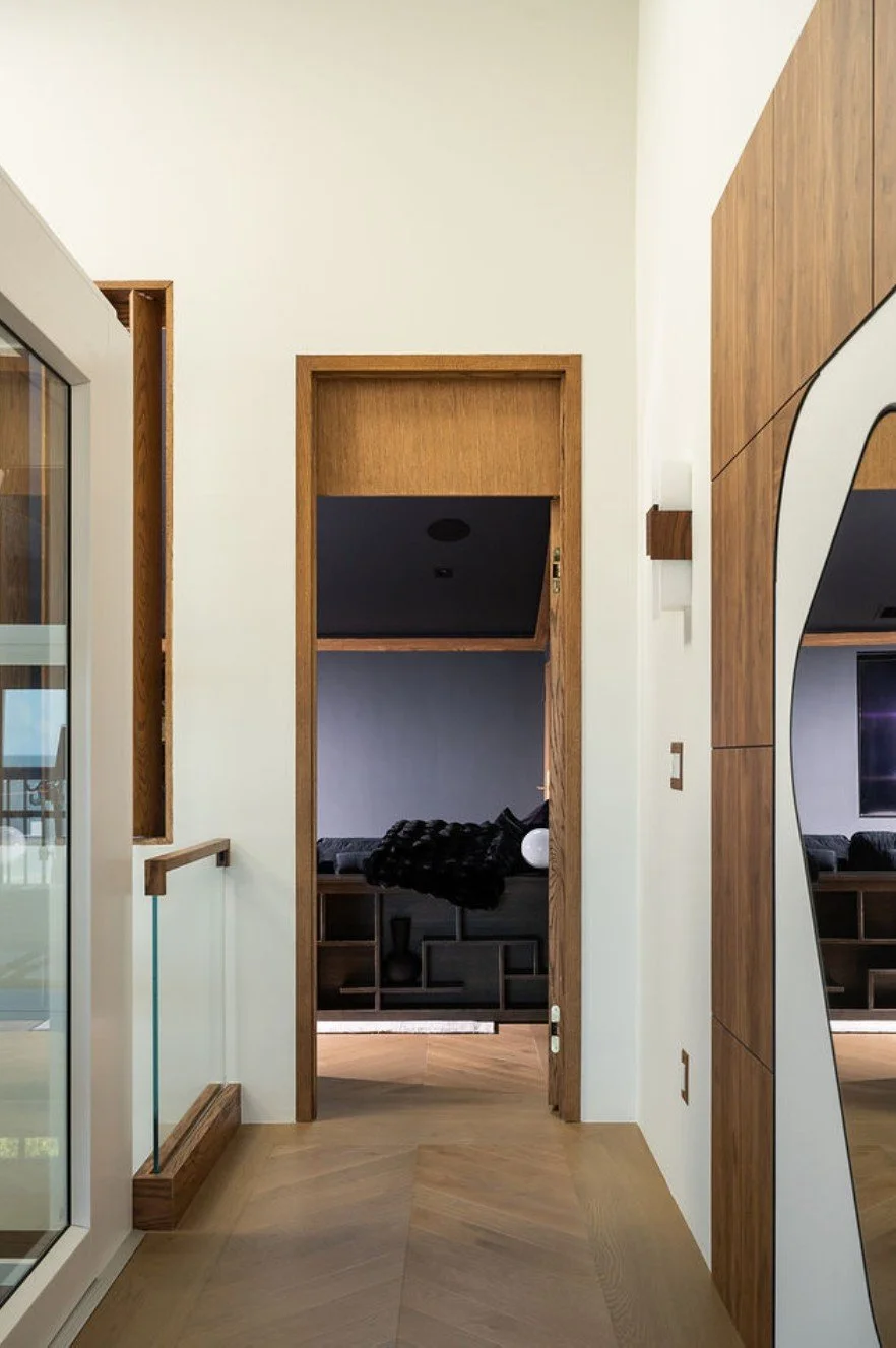 Interior view of a modern home hallway with a wooden door frame leading to a living room with a black sofa and a black blanket, white walls, wood paneling, and a large mirror on the right.