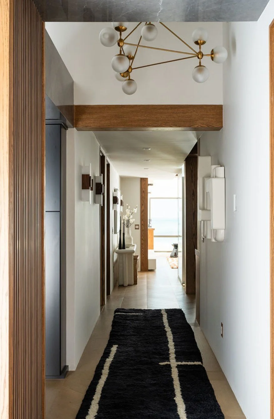 Interior view of a modern hallway with a black and white runner rug, white walls, wooden accents, a contemporary chandelier, and sunlight coming through a large window at the end.