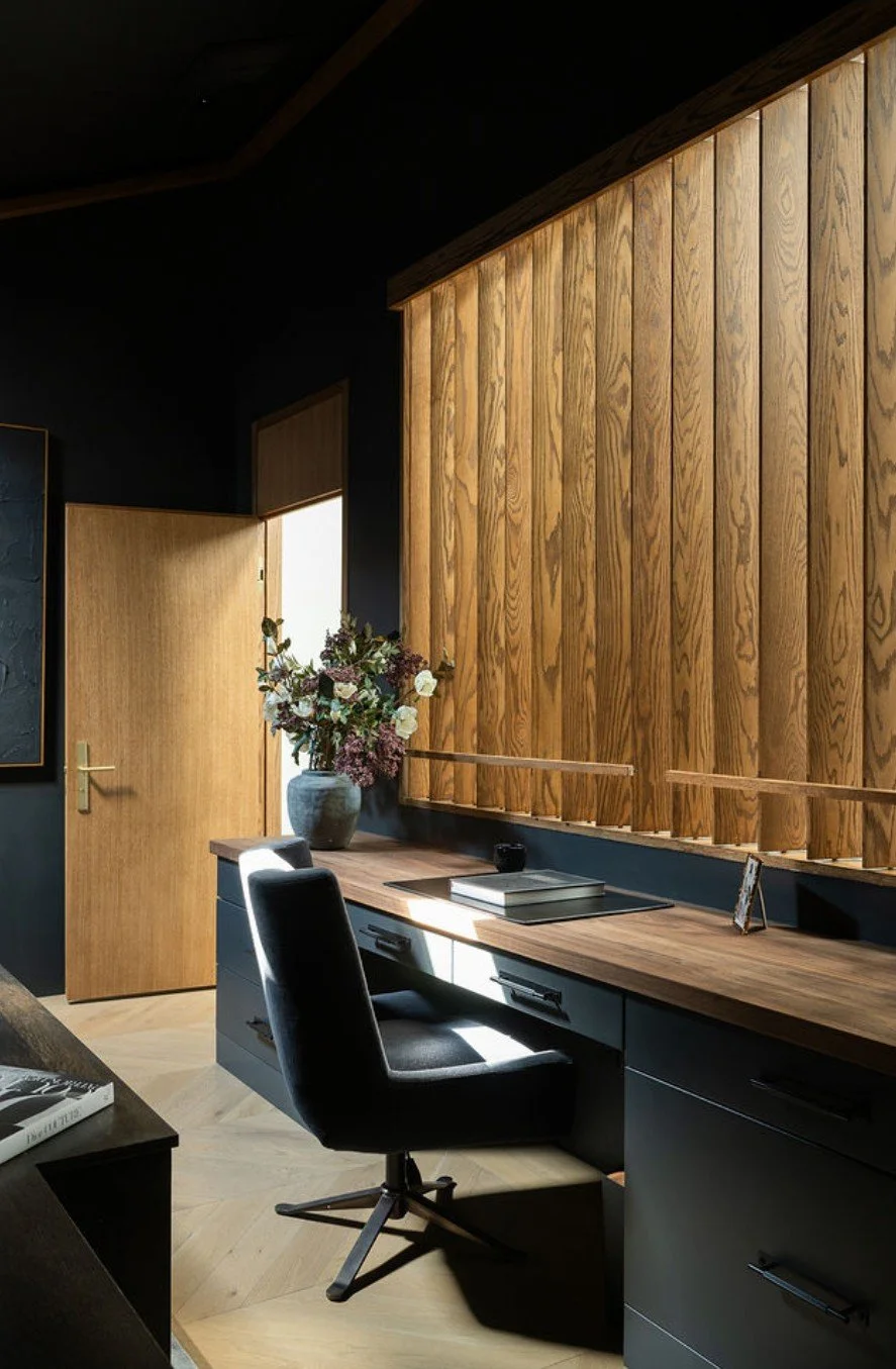 Modern home office with a black desk, a black swivel chair, a wooden wall panel, a potted plant, and a door with natural light streaming in.