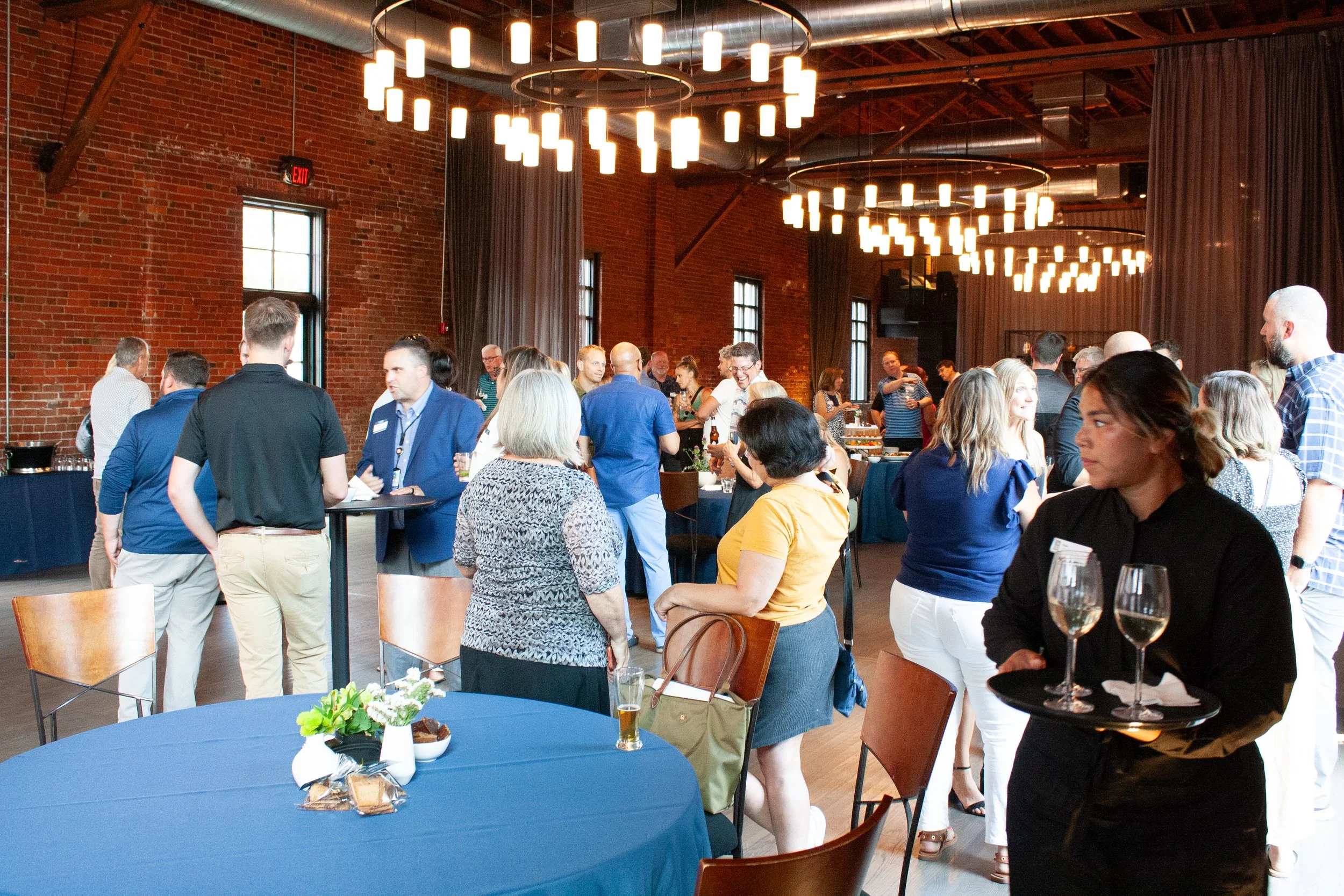A business happy hour party is in full swing at High Line Car House, an industrial event venue in Columbus, Ohio. An event server is in the foreground collecting wineglasses. 