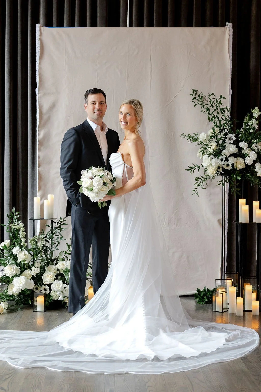 A couple stands, smiling at the camera in front of their ceremony altar at a historic wedding venue in Columbus, Ohio. There are candles and dramatic white flower arrangements on either side of them. Behind the newlyweds is a flat fabric backdrop.