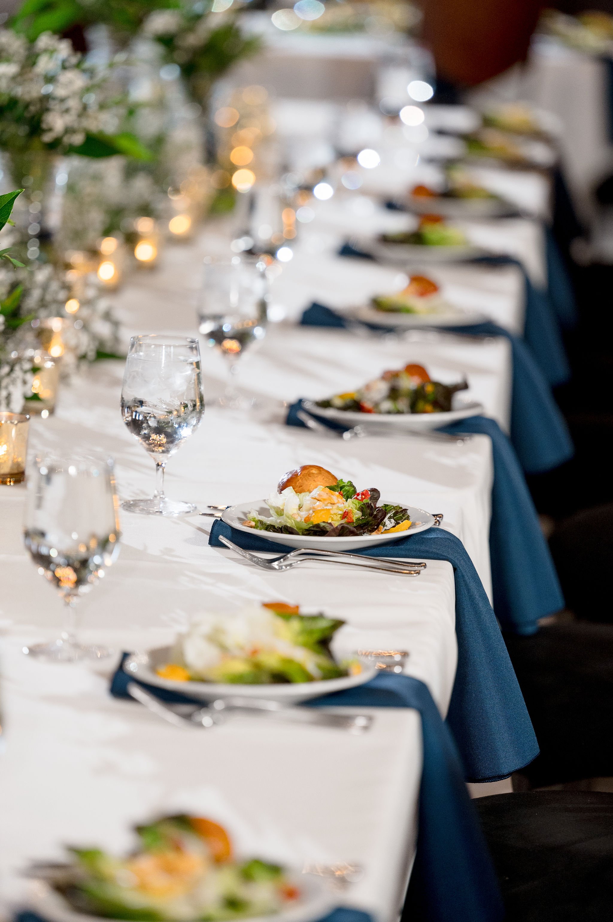  A long white linen table is set for salad service. Each place setting has a glass of water, a blue napkin, and flatware.  