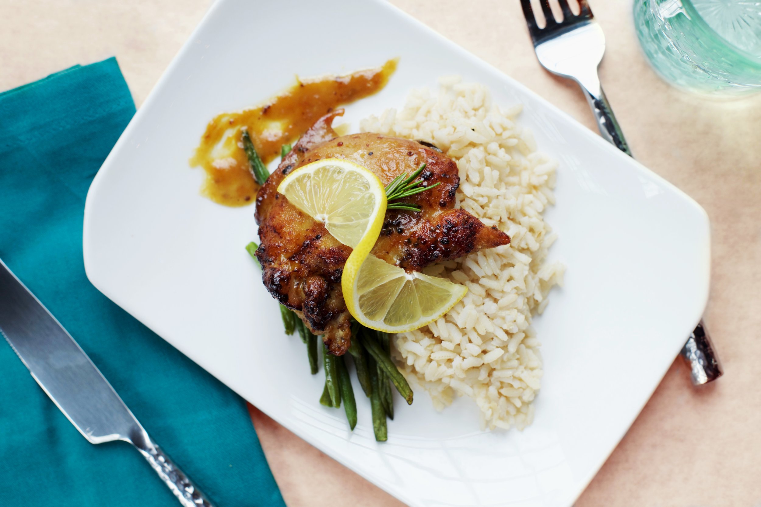  A roasted chicken is plated atop a bed of rice and green beans, garnished with a twisted lemon. The plate is framed by a fork and a knife and a blue napkin. 